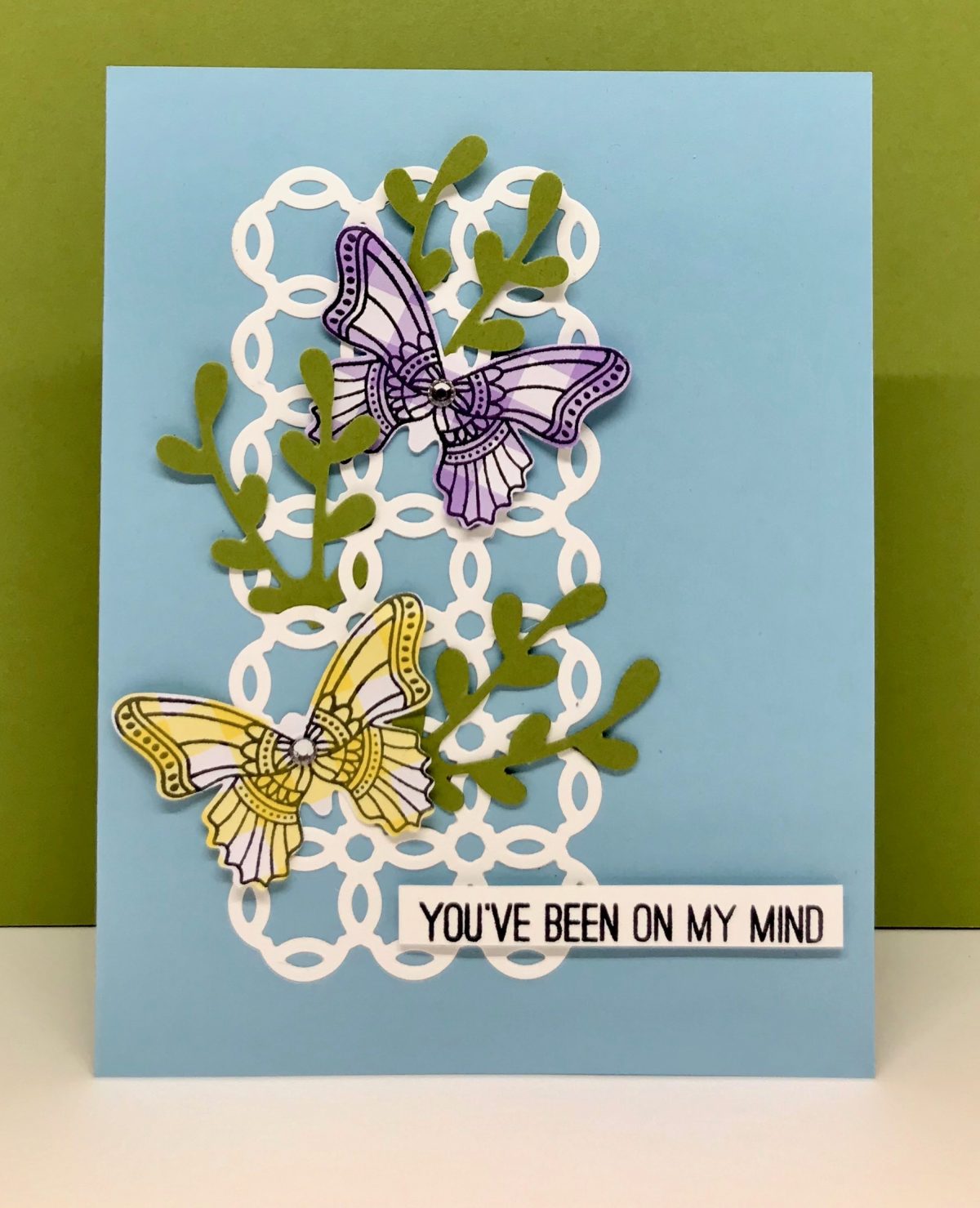 A card with punched out butterflies stamped on pastel gingham checked designer paper on white lattice on blue background 